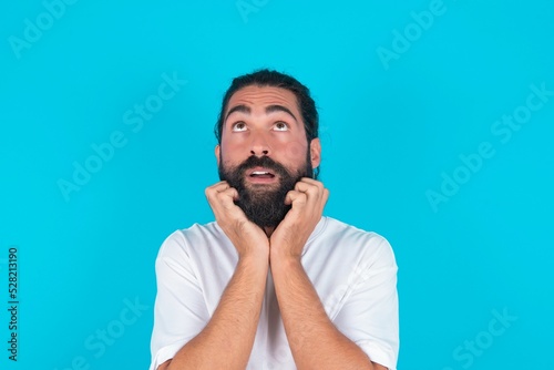 Portrait of sad young bearded man wearing white T-shirt over blue studio background hands face look empty space