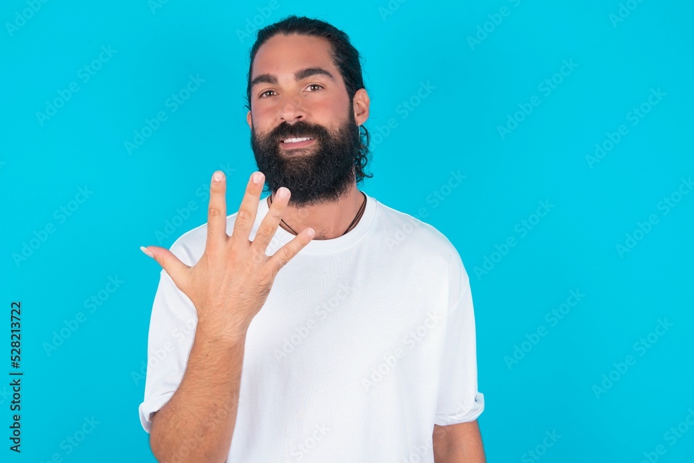 Naklejka premium young bearded man wearing white T-shirt over blue studio background smiling and looking friendly, showing number five or fifth with hand forward, counting down