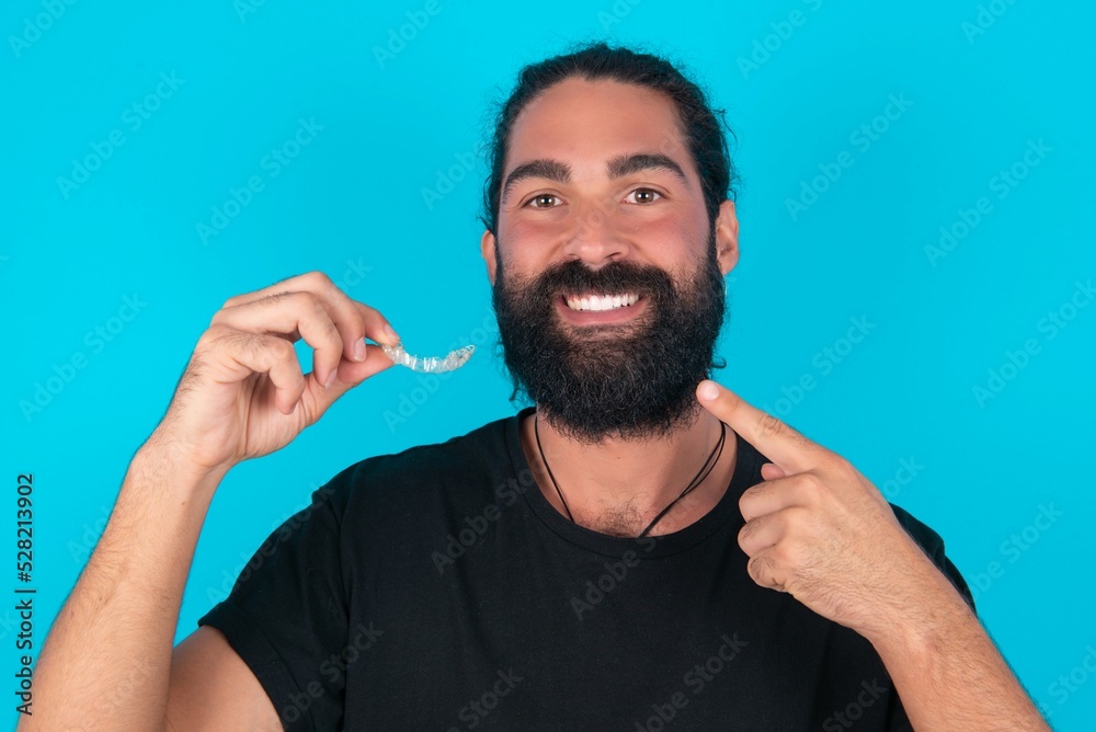 young bearded man wearing black T-shirt over blue studio background holding an invisible aligner and pointing to her perfect straight teeth. Dental healthcare and confidence concept. young bearded man wearing black T-shirt over blue studio background holding an invisible aligner and pointing to her perfect straight teeth. Dental healthcare and confidence concept.