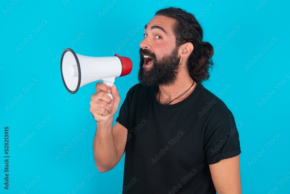 young bearded man wearing black T-shirt over blue studio background Through Megaphone with Available Copy Space young bearded man wearing black T-shirt over blue studio background Through Megaphone with Available Copy Space
