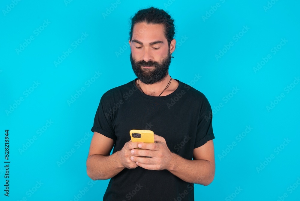 Excited young bearded man wearing black T-shirt over blue studio background winking and eye hold smart phone use read social network news Excited young bearded man wearing black T-shirt over blue studio background winking and eye hold smart phone use read social network news