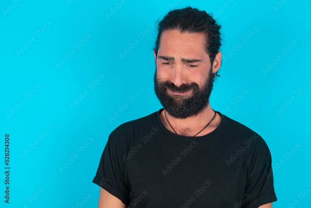Obraz premium Displeased upset young bearded man wearing black T-shirt over blue studio background frowns face as going to cry, being discontent and unhappy as can't achieve goals, Disappointed model has troubles