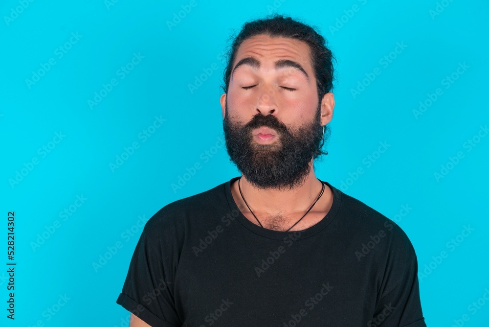 Shot of pleasant looking young bearded man wearing black T-shirt over blue studio background, pouts lips, looks at camera, Human facial expressions Shot of pleasant looking young bearded man wearing black T-shirt over blue studio background, pouts lips, looks at camera, Human facial expressions