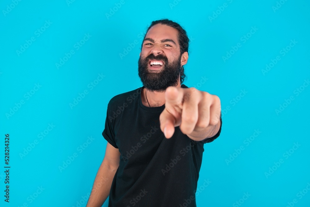 Naklejka premium young bearded man wearing black T-shirt over blue studio background pointing displeased and frustrated to the camera, angry and furious ready to fight with you.