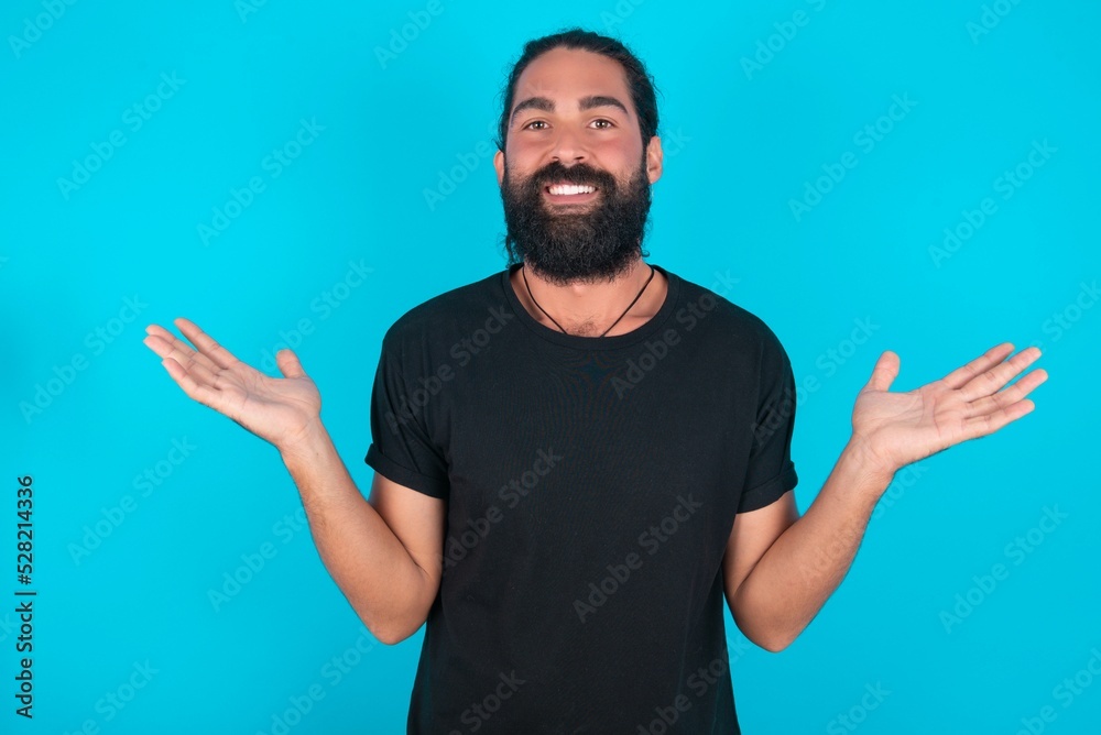 Cheerful cheery optimistic young bearded man wearing black T-shirt over blue studio background holding two palms copy space Cheerful cheery optimistic young bearded man wearing black T-shirt over blue studio background holding two palms copy space