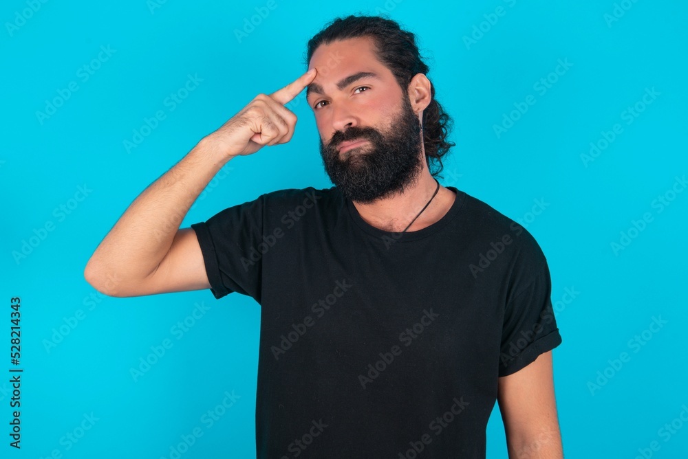 young bearded man wearing black T-shirt over blue studio background pointing unhappy at pimple on forehead, blackhead infection. Skincare concept. young bearded man wearing black T-shirt over blue studio background pointing unhappy at pimple on forehead, blackhead infection. Skincare concept.