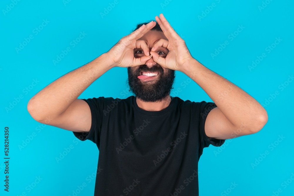young bearded man wearing black T-shirt over blue studio background doing ok gesture like binoculars sticking tongue out, eyes looking through fingers. Crazy expression. young bearded man wearing black T-shirt over blue studio background doing ok gesture like binoculars sticking tongue out, eyes looking through fingers. Crazy expression.