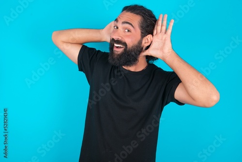 young bearded man wearing black T-shirt over blue studio background Trying to hear both hands on ear gesture, curious for gossip. Hearing problem, deaf