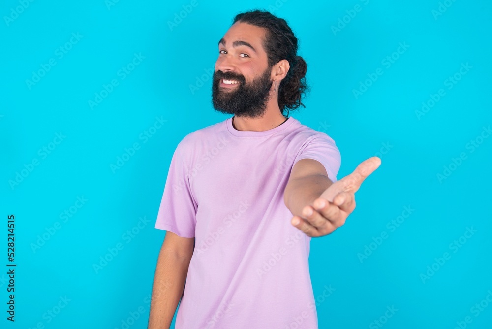young bearded man wearing violet T-shirt over blue studio background smiling friendly offering handshake as greeting and welcoming. Successful business.