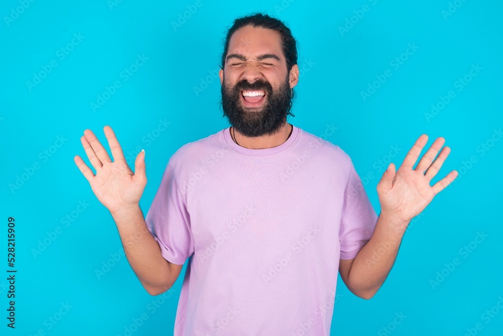 Emotive young bearded man wearing violet T-shirt over blue studio background laughs loudly, hears funny joke or story, raises palms with satisfaction, Emotive young bearded man wearing violet T-shirt over blue studio background laughs loudly, hears funny joke or story, raises palms with satisfaction,