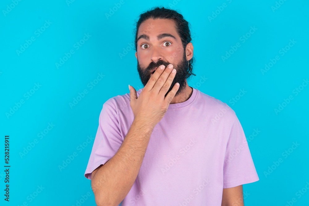 Emotional young bearded man wearing violet T-shirt over blue studio background gasps from astonishment, covers opened mouth with palm, looks shocked at camera. Emotional young bearded man wearing violet T-shirt over blue studio background gasps from astonishment, covers opened mouth with palm, looks shocked at camera.