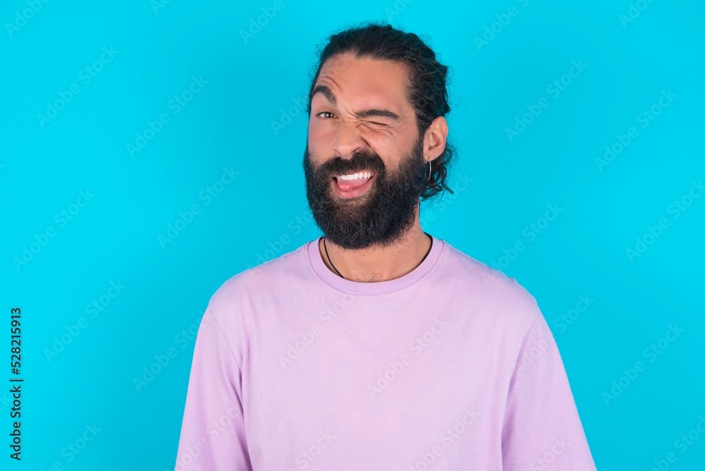 Coquettish young bearded man wearing violet T-shirt over blue studio background smiling happily, blinking at camera in a playful manner, flirting with you.