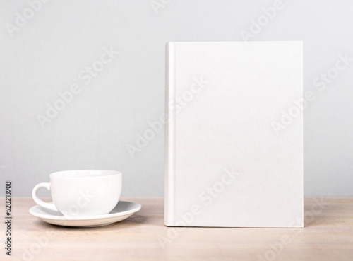 Book mockup and cup at wooden table. Reading leisure, relaxation with tea or coffee mug, harmony concept. Novel, encyclopedia, code template with empty cover.