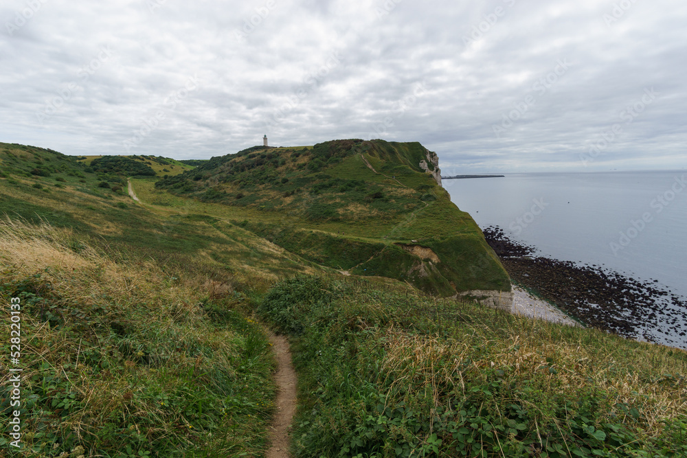 Coastal hiking trail at the cliffs of the Alabaster coast through green meadows with distant lighthouse Phare d'Antifer, Etretat, Normandy, France Coastal hiking trail at the cliffs of the Alabaster coast through green meadows with distant lighthouse Phare d'Antifer, Etretat, Normandy, France