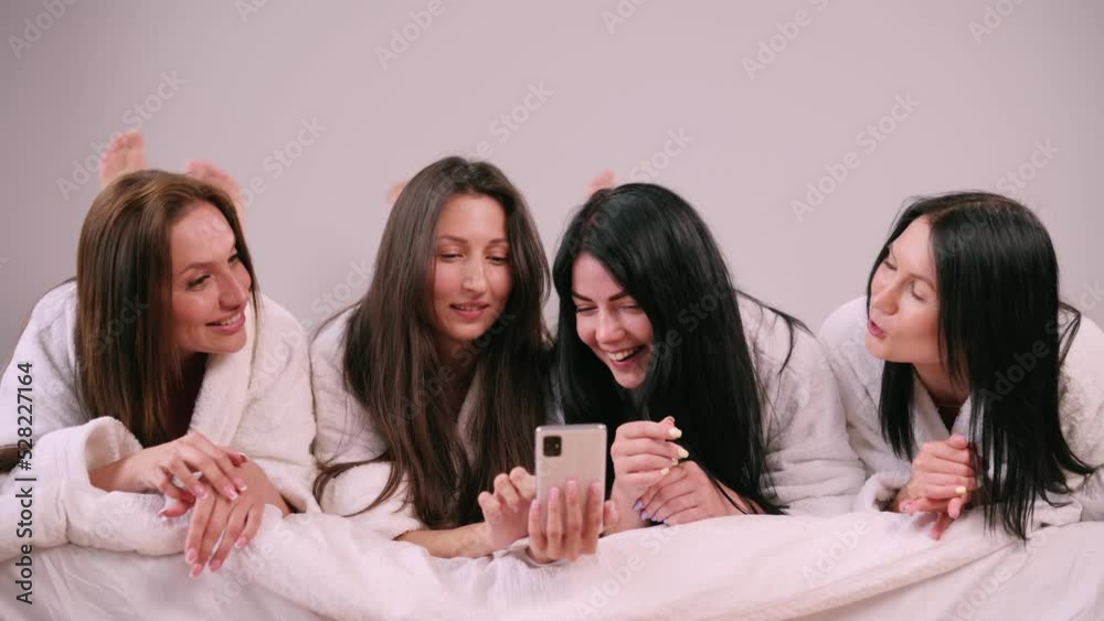 Girlfriends have fun chatting together. Young attractive females are lying in bed looking at the smartphone screen and laughing. Girlfriends have fun chatting together. Young attractive females are lying in bed looking at the smartphone screen and laughing.