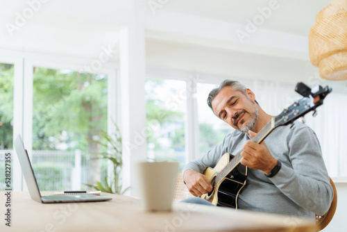 Smiling bearded musician Mature Adult Man playing acoustic guitar, Using Laptop