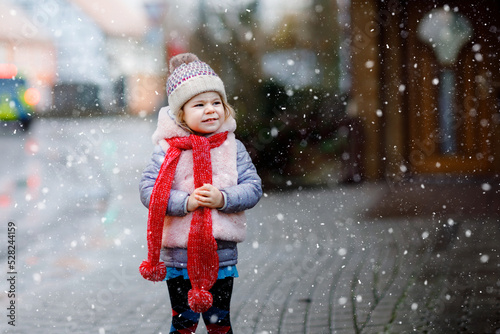 Cute little funny toddler girl in colorful winter fashion clothes having fun and playing with snow, outdoors during snowfall. Active outdoors leisure with children. Happy healthy child