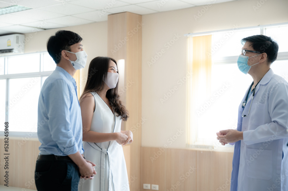 Young male doctor wearing covid-19 safety mask in conversation with young couple in clinic and hospital passage Young male doctor wearing covid-19 safety mask in conversation with young couple in clinic and hospital passage