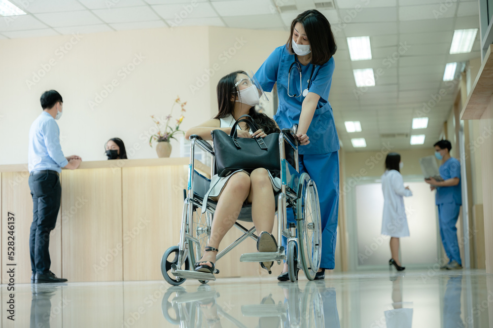 Smiling young female doctor in scrub and covid-19 face mask in conversation with patient sitting in wheelchair holding purse during discharge from hospital Smiling young female doctor in scrub and covid-19 face mask in conversation with patient sitting in wheelchair holding purse during discharge from hospital