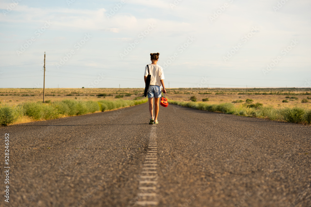 Obraz premium A girl walks along an empty road in the steppe. She has a ukulele in her hands and a backpack on her shoulders. It's summer and warm outside. Open road.