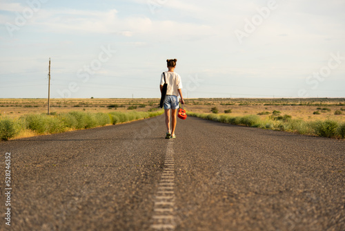 A girl walks along an empty road in the steppe. She has a ukulele in her hands and a backpack on her shoulders. It s summer and warm outside. Open road.