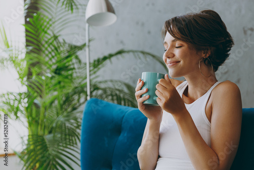 Cuadro en lienzo Close up side view young woman wear white tank shirt drink tea coffee sniff sit on blue sofa stay at home flat rest relax spend free spare time in living room indoors grey wall People lounge concept