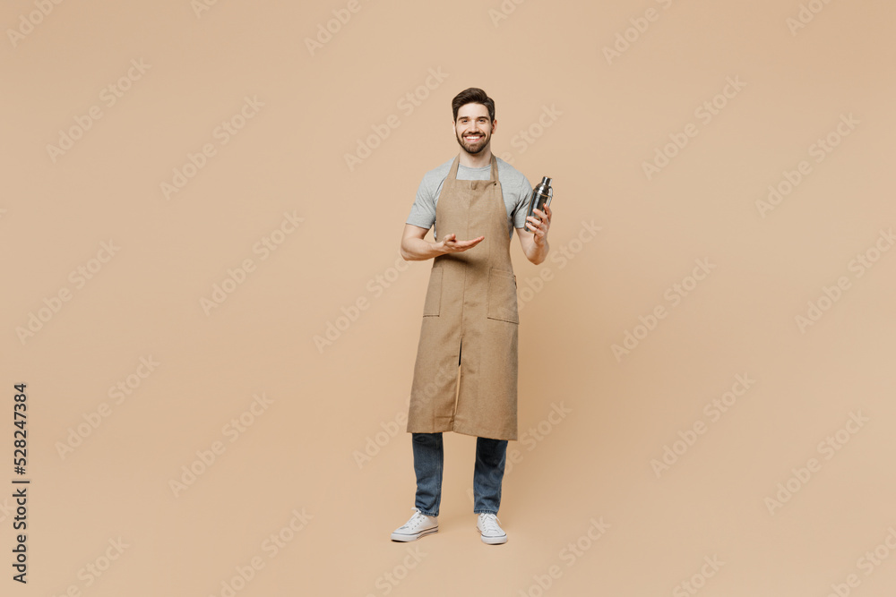 Full body young man barista barman employee wear brown apron work in bar pub nightclub hold shaker make alcohol cocktail isolated on plain pastel light beige background Small business startup concept. Full body young man barista barman employee wear brown apron work in bar pub nightclub hold shaker make alcohol cocktail isolated on plain pastel light beige background Small business startup concept.