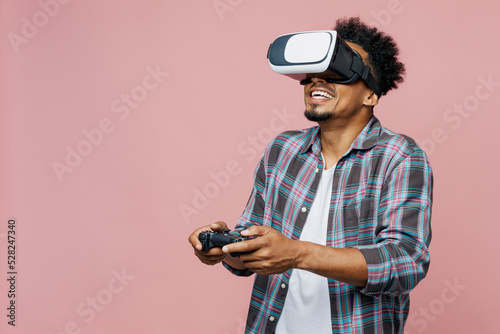 Young gambling man of African American ethnicity 20s he wear blue shirt hold in hand play pc game with joystick console watching in vr headset pc gadget isolated on plain pastel light pink background.