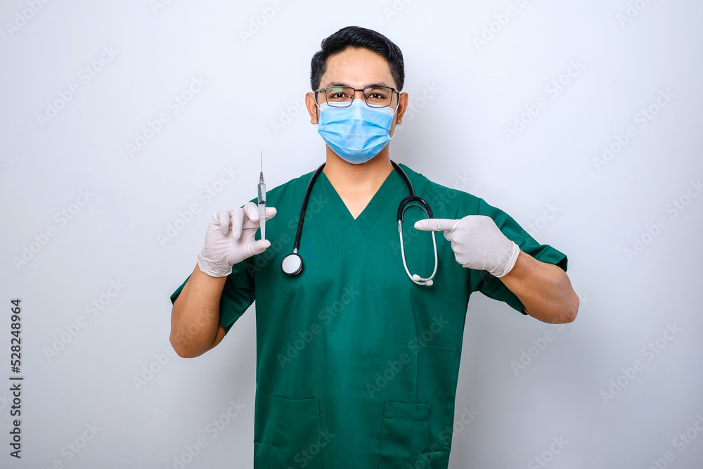 Serious male nurse or doctor in green scrubs, medical mask, pointing at syringe with coronavirus vaccine Serious male nurse or doctor in green scrubs, medical mask, pointing at syringe with coronavirus vaccine