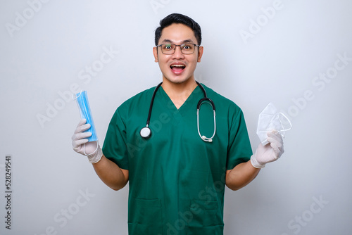 Cheerful smiling asian male doctor in scrubs holding medical mask but suggest respirator, handing in to patient