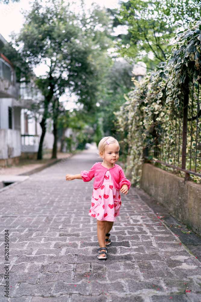 Little girl walks along the paving stones past the wrought-iron fence of the park. High quality photo