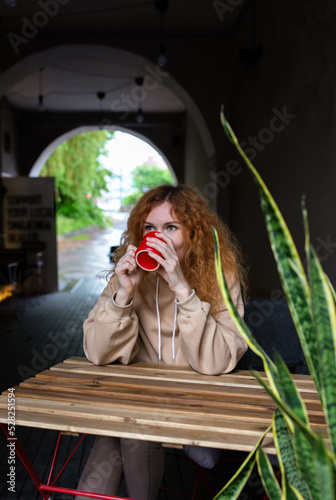  Young red-haired woman alone drinking a drink in a terrace cafe on a rainy day