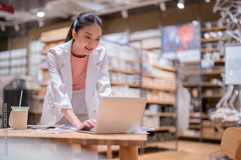 Fototapeta premium asian female smart entrepreneur working use laptop device stock manage and checking product inventory in warehouse showroom,asia woman smiling cheerful business owner work in showroom warehouse store