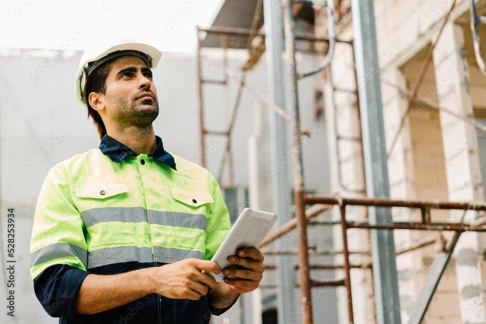 Construction worker with safety hardhat and in vest looking at tablet computer on construction site Construction worker with safety hardhat and in vest looking at tablet computer on construction site