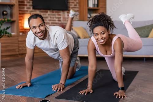Glad young african american male and female in sportswear doing leg exercises on mat in living room