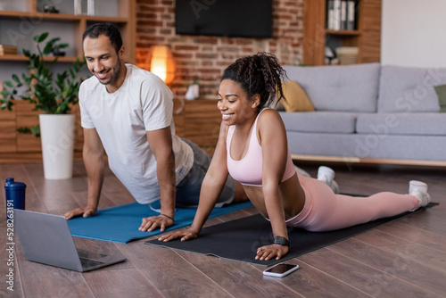 Glad young black couple in sportswear do stretching or practicing yoga, watch video lesson on laptop on mat