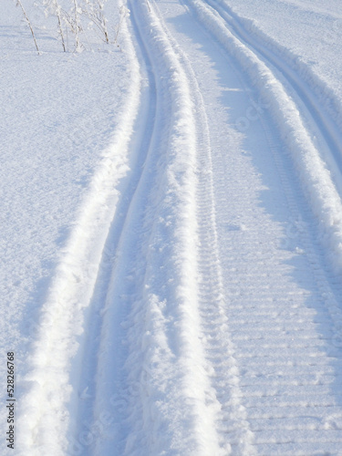 Track of traces from a snowmobile in drifts of white snow. Nature and outdoor on a winter sunny day. Vertical illustration with snowy field © Deacon docs