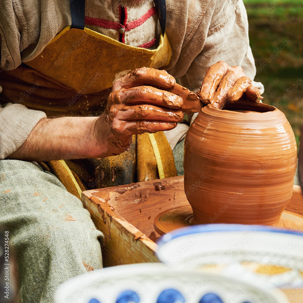 A man in old medieval Byzantine clothes sits behind a vintage potter's wheel and makes dishes out of clay. Pottery in nature in retro style.