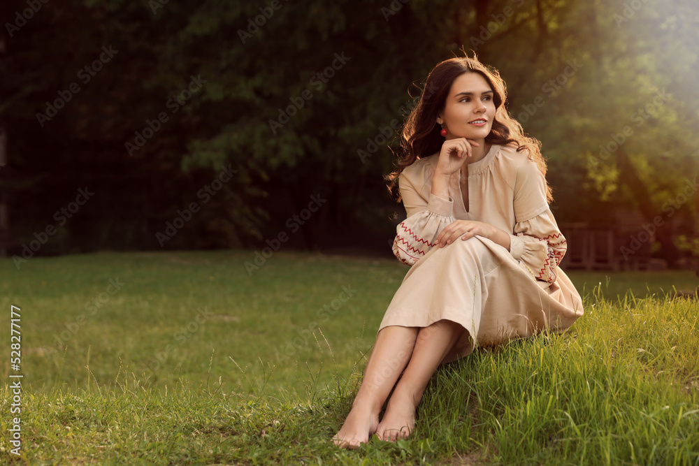 Beautiful woman in embroidered dress sitting on green grass outdoors, space for text. Ukrainian national clothes Beautiful woman in embroidered dress sitting on green grass outdoors, space for text. Ukrainian national clothes