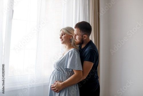 A man hugs his pregnant wife from behind and touches his belly near the window of the house. Pregnant couple. Lifestyle. 