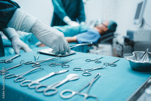 Close up surgeon doctor's hand with hygiene glove taking sterile surgical instrument tool, equipment in operating room patient in background for surgery. Hospital, medical healthcare emergency concept