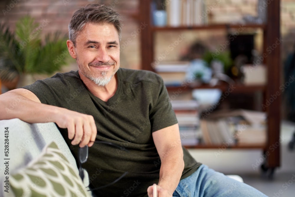 Portrait of happy handsome mature man with beard looking at camera, sitting on sofa at home in the living room.