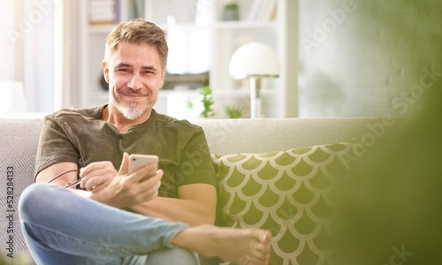Portrait of middle aged handsome man wearing casual clothes, sitting on sofa at home in the living room, using phone, looking at camera.