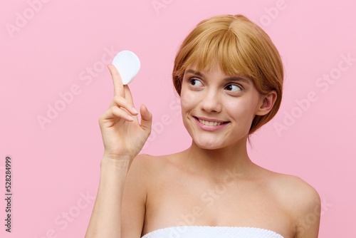 horizontal photo of a happy, sophisticated woman with blonde hair gathered back, standing on a pink background, holding a cotton pad in her hand and smiling sweetly at him