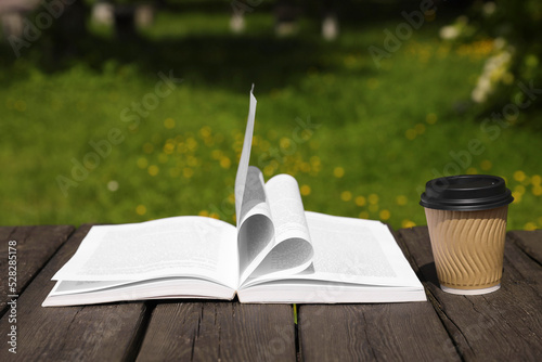 Open book and paper coffee cup on wooden table outdoors