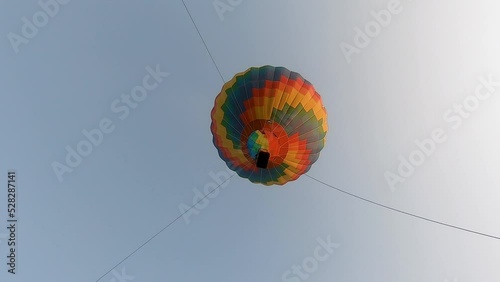  Bottom-up view of a flying multicolored balloon with a basket for passengers, tied with a rope to the ground, against a blue sky background  
