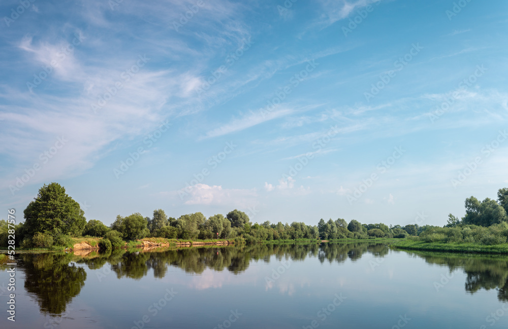 Summer landscape. Calm Berezina river with green banks under a blue sky with clouds.