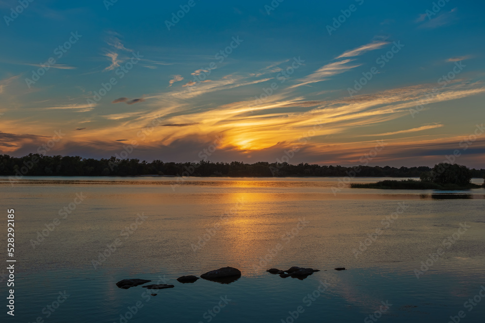 Beautiful sunset over the Dnieper River. Yellow cirrus clouds over the forest. Sun light reflection and stones in water