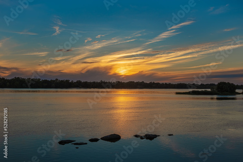 Beautiful sunset over the Dnieper River. Yellow cirrus clouds over the forest. Sun light reflection and stones in water