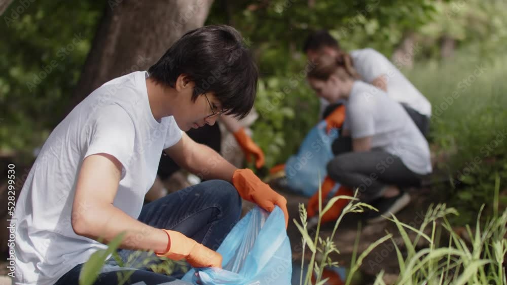 Group of eco-activists removes garbage in nature, an Asian man puts garbage in a plastic bag, looks at the camera and smiles. Green life concept Group of eco-activists removes garbage in nature, an Asian man puts garbage in a plastic bag, looks at the camera and smiles. Green life concept